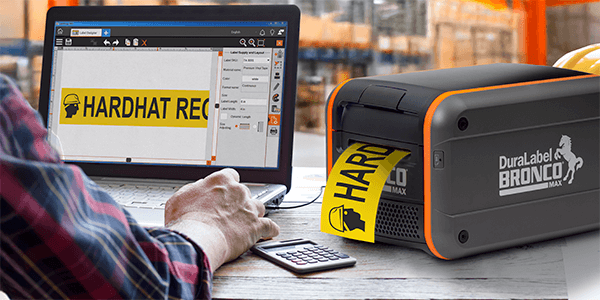 DuraLabel Bronco Max printer producing a yellow “Hardhat Required” label beside a laptop.